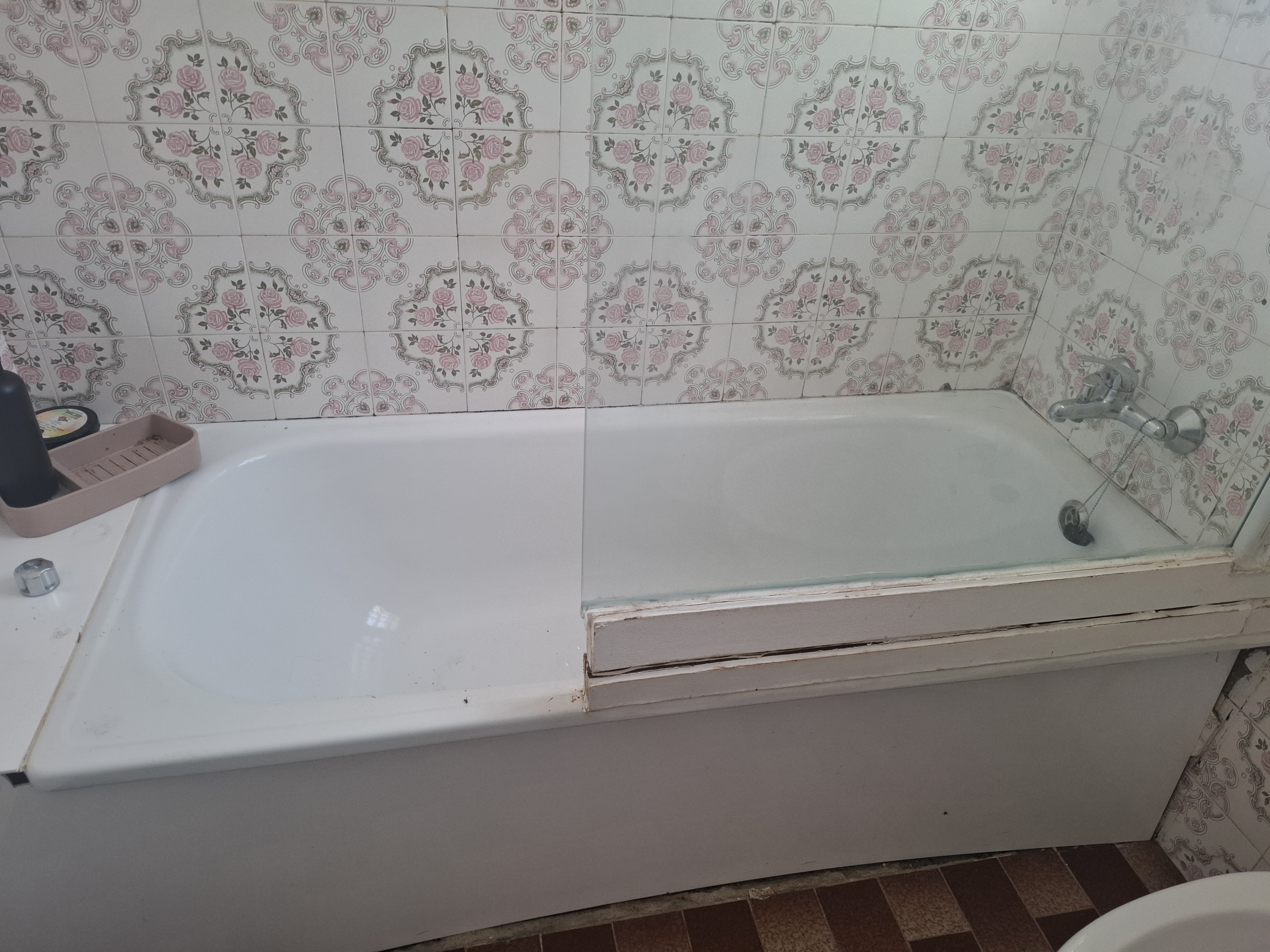 White bathtub against a wall covered in pink floral patterned tiles; chrome faucet visible on the right and a pink soap dish on the edge.