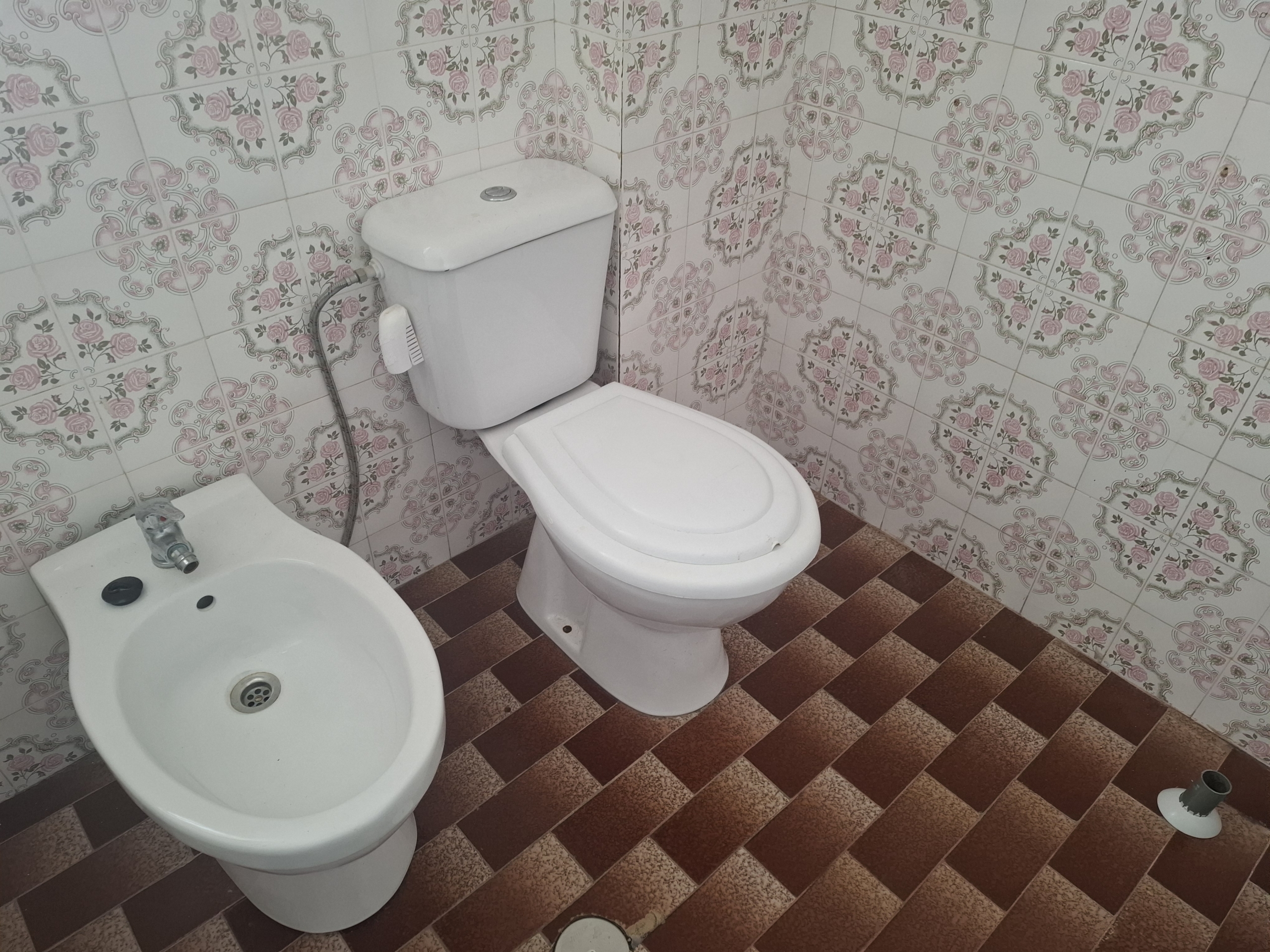 White toilet beside a bidet in a bathroom with pink floral patterned wall tiles and brown tiled floor