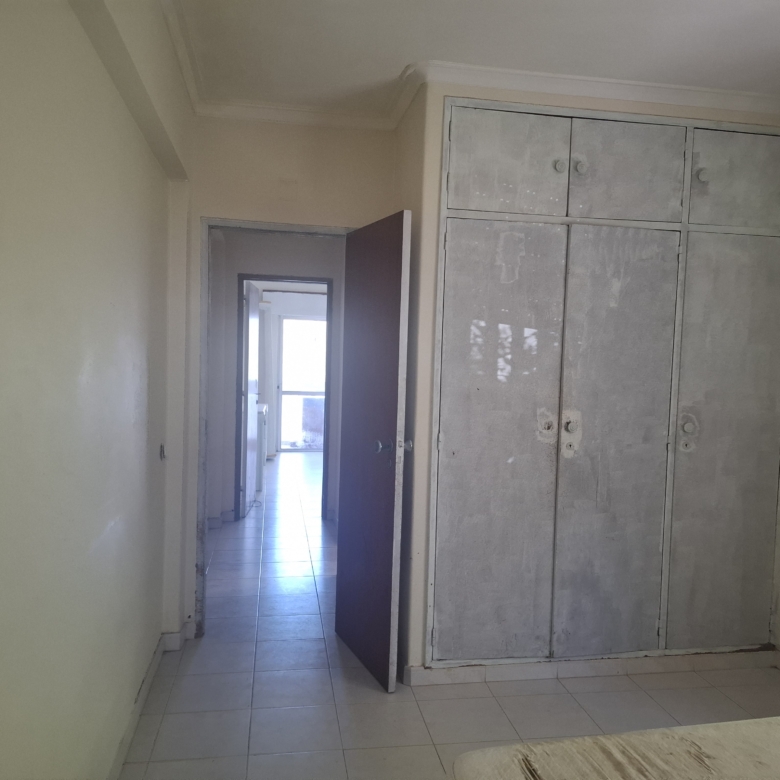 Narrow hallway with pale yellow walls, a tall gray metal wardrobe along the right wall, and an open door at the far end leading to a sunlit room with tiled flooring.