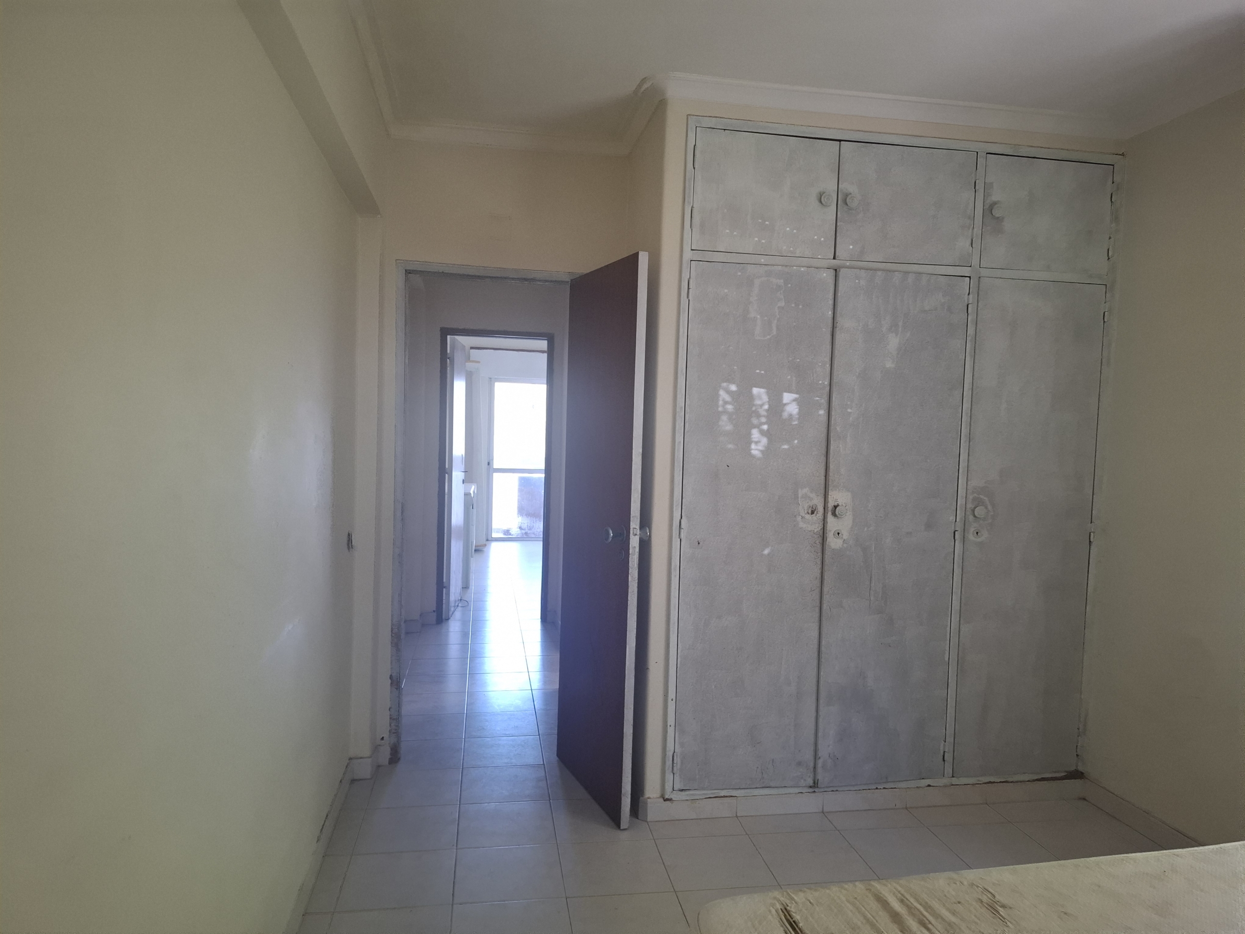 Narrow hallway with pale yellow walls, a tall gray metal wardrobe along the right wall, and an open door at the far end leading to a sunlit room with tiled flooring.