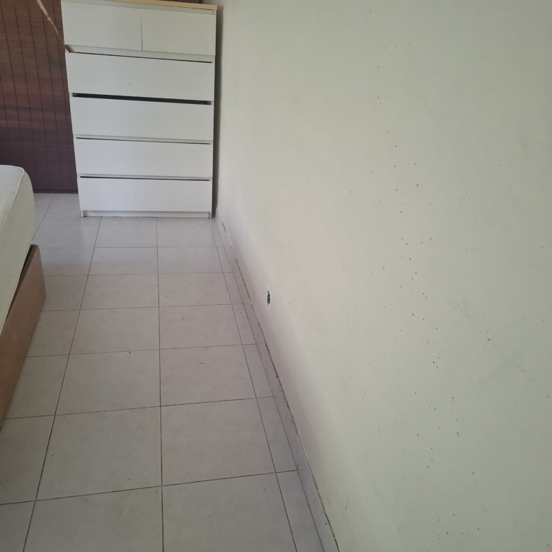 Corner of a sparse bedroom with a white 5-drawer dresser beside a bed and a pale green wall with small marks beyond.