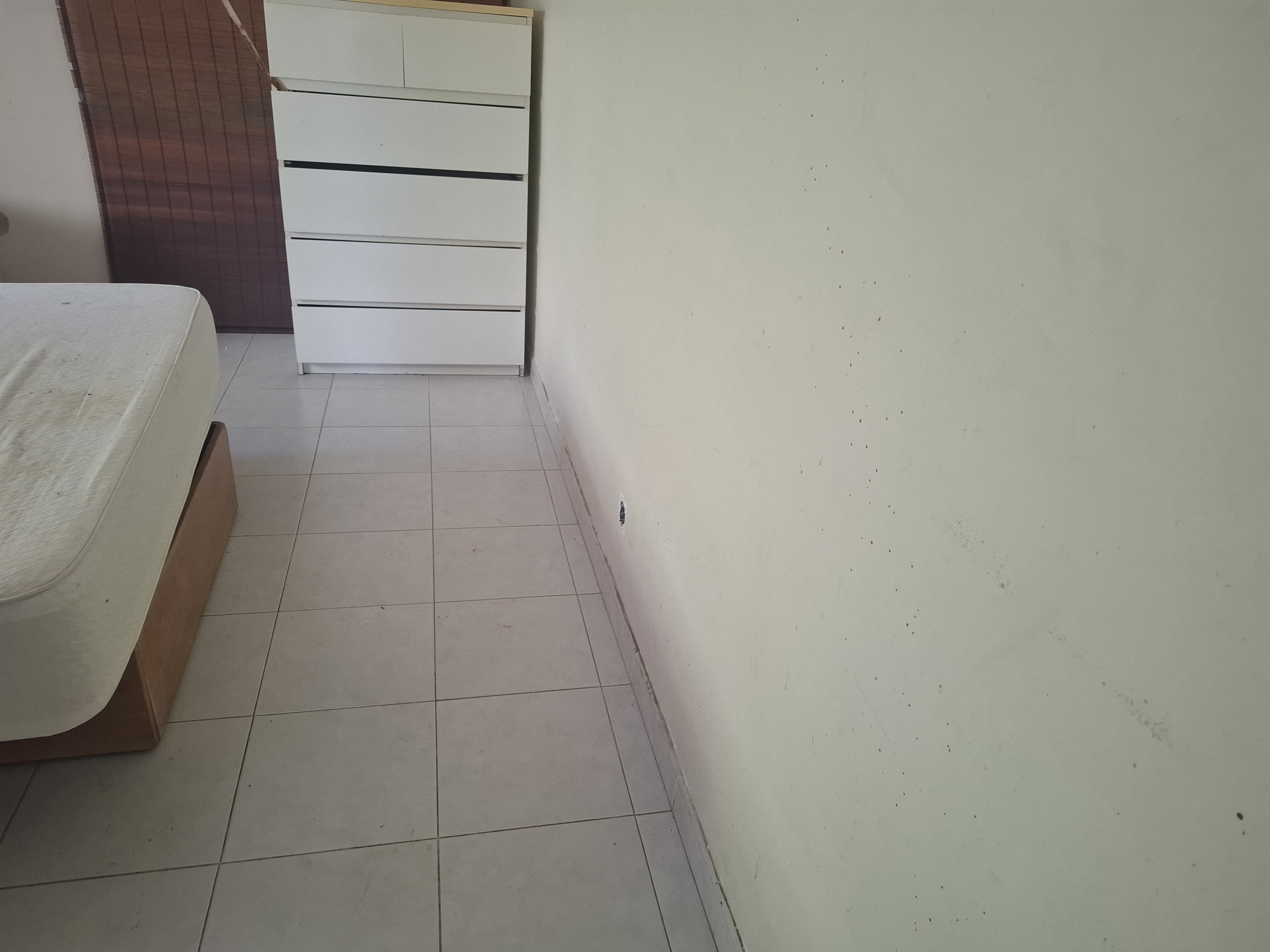 Corner of a sparse bedroom with a white 5-drawer dresser beside a bed and a pale green wall with small marks beyond.