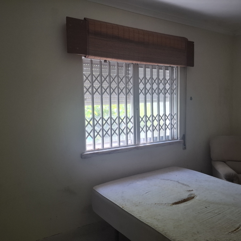 Dimly lit room with a barred window and wooden horizontal blind, a chipped table in the foreground, and a beige sofa in the corner.