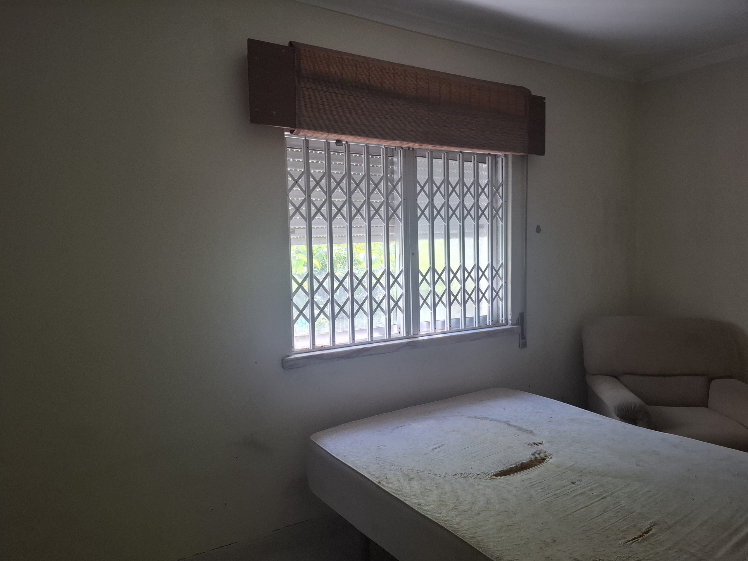 Dimly lit room with a barred window and wooden horizontal blind, a chipped table in the foreground, and a beige sofa in the corner.