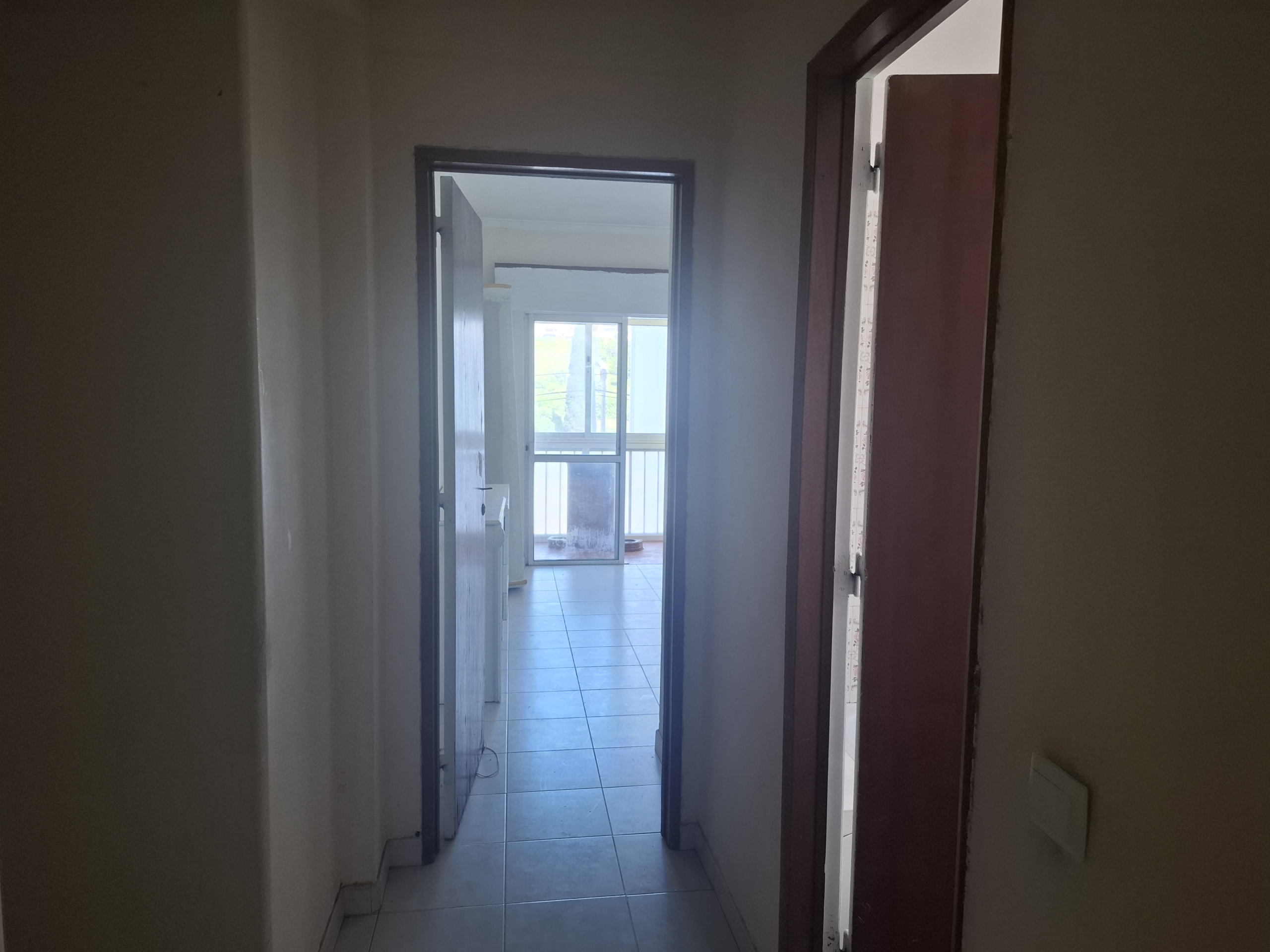 Narrow hallway with open doorways opening to a bright room with large windows and a balcony outside the doors.