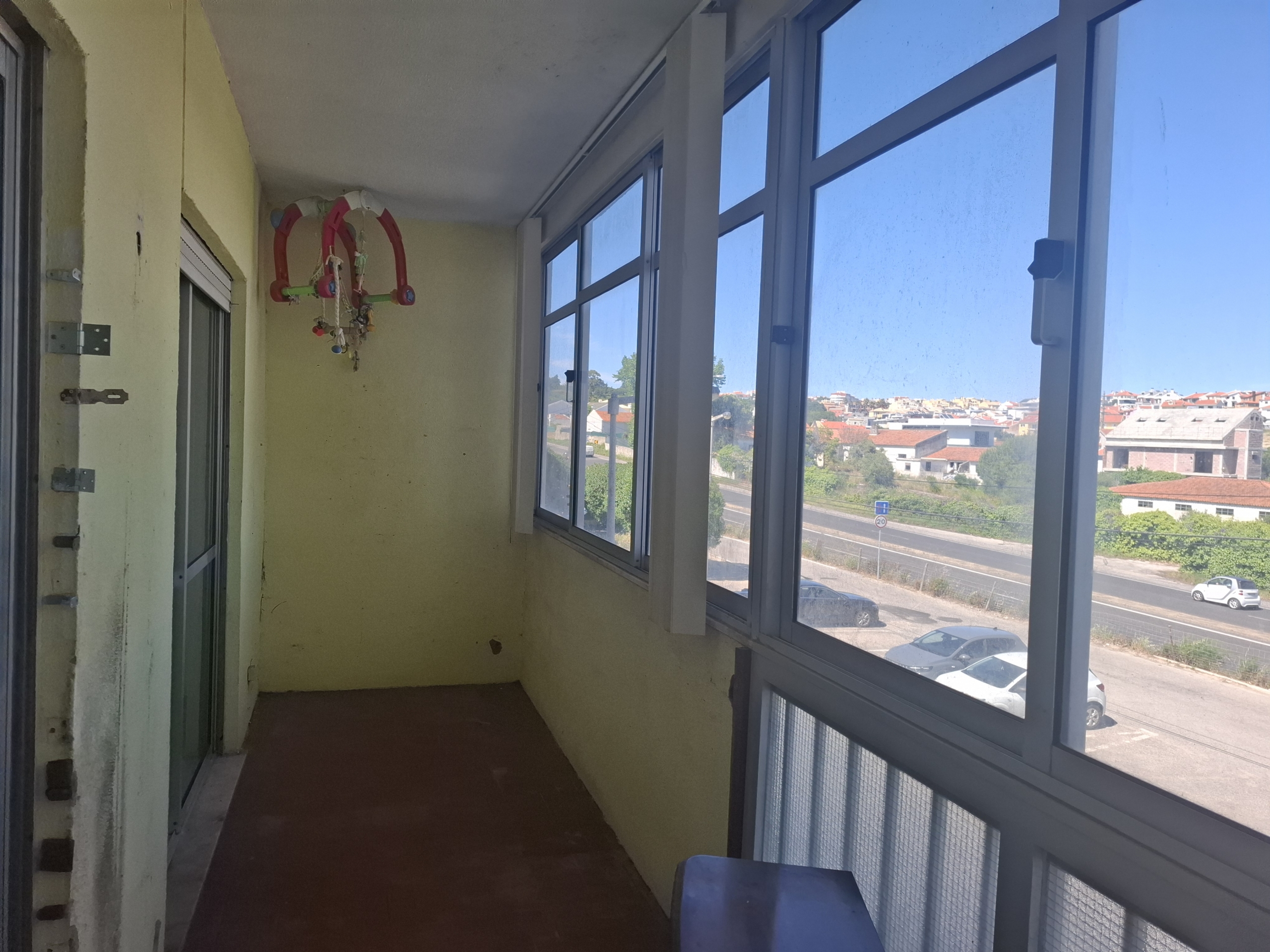 Narrow corridor balcony with large windows on the right, yellow walls, and a colorful hanging toy or mobile near the far wall on the left side.