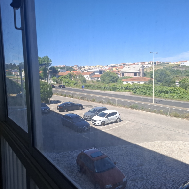 View from an open window onto a parking lot with several cars and a hillside town in the distance on a bright day.