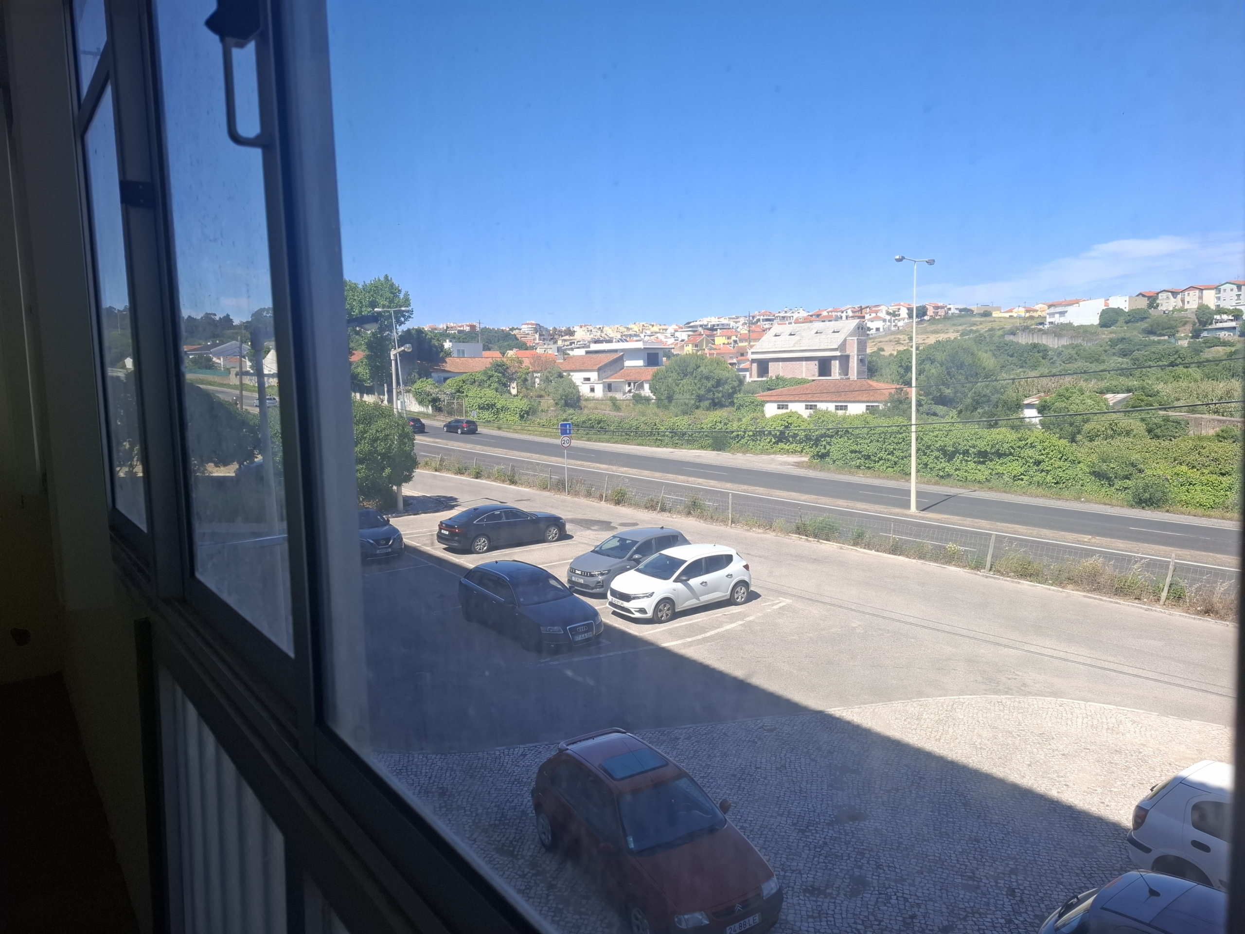 View from an open window onto a parking lot with several cars and a hillside town in the distance on a bright day.