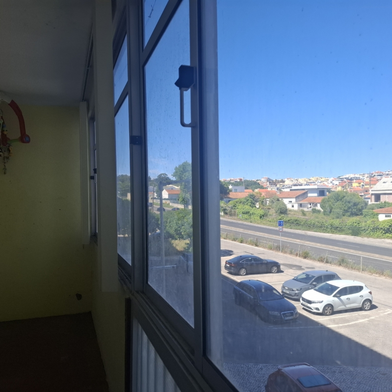 Interior view of a yellow-walled room with a colorful baby mobile by a large window; outside, a parking lot and hillside houses under a sunny sky,