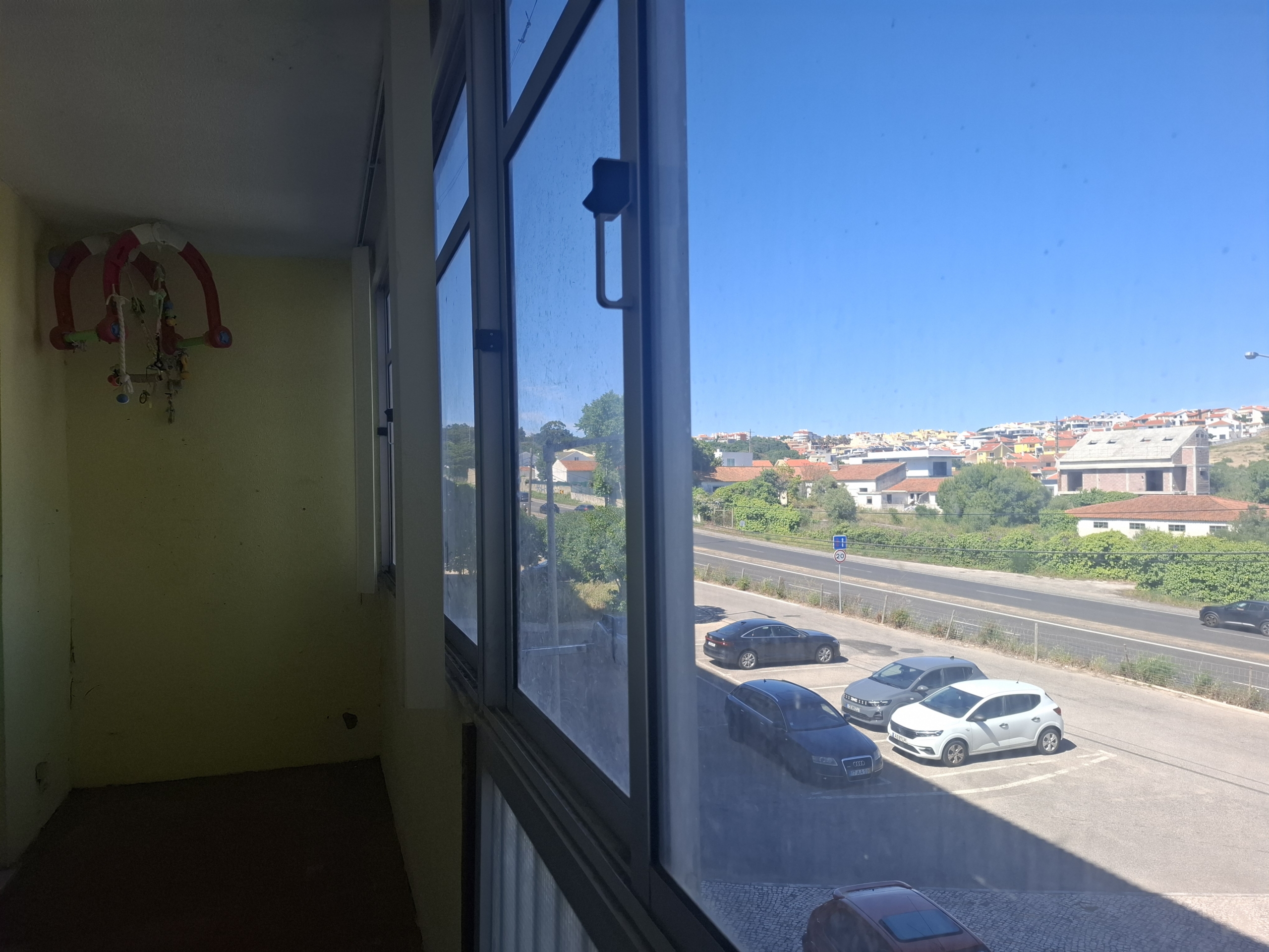 Interior view of a yellow-walled room with a colorful baby mobile by a large window; outside, a parking lot and hillside houses under a sunny sky,