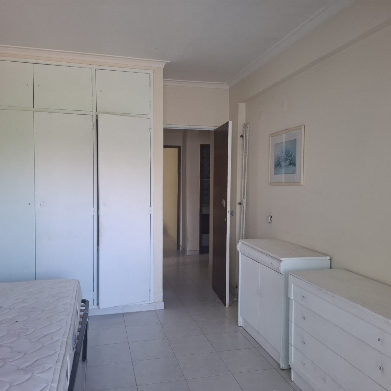 Bedroom with tall white built-in wardrobe along left wall, and white dressers; doorway to hallway visible in center-right; neutral walls and framed art.