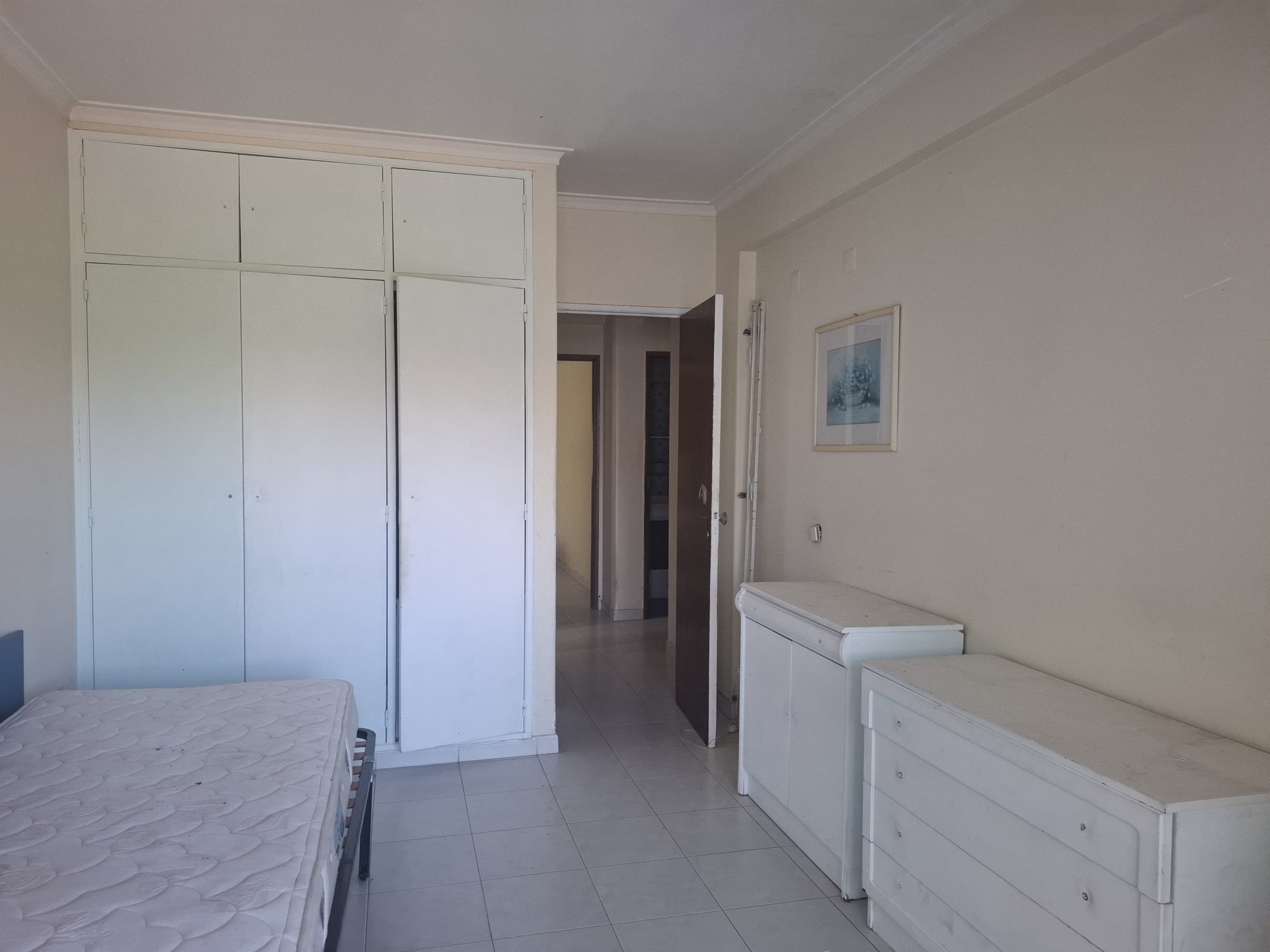 Bedroom with tall white built-in wardrobe along left wall, and white dressers; doorway to hallway visible in center-right; neutral walls and framed art.