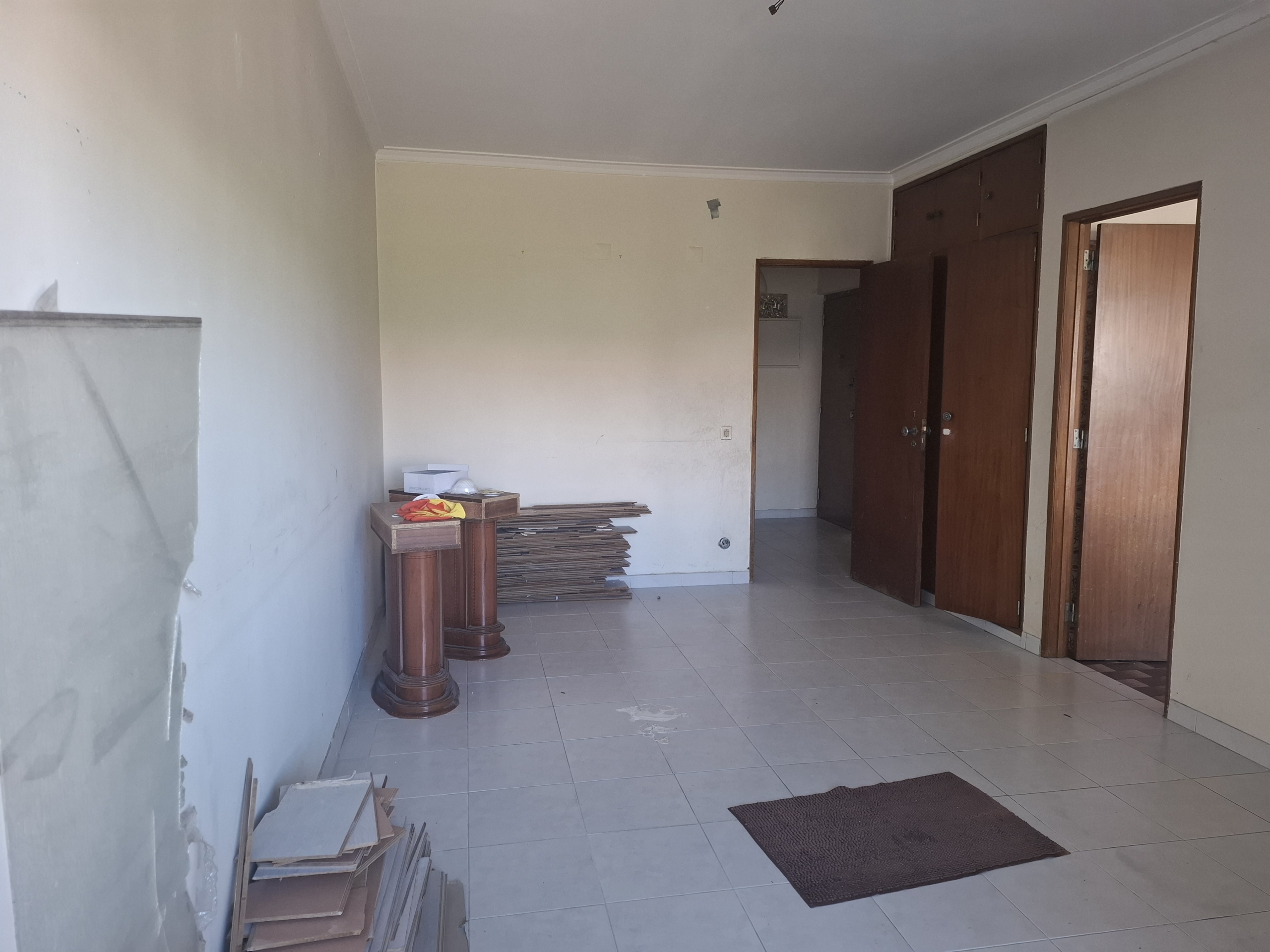 Empty room under renovation with piles of wooden planks, a small pedestal table, beige walls, and open brown cabinet doors.