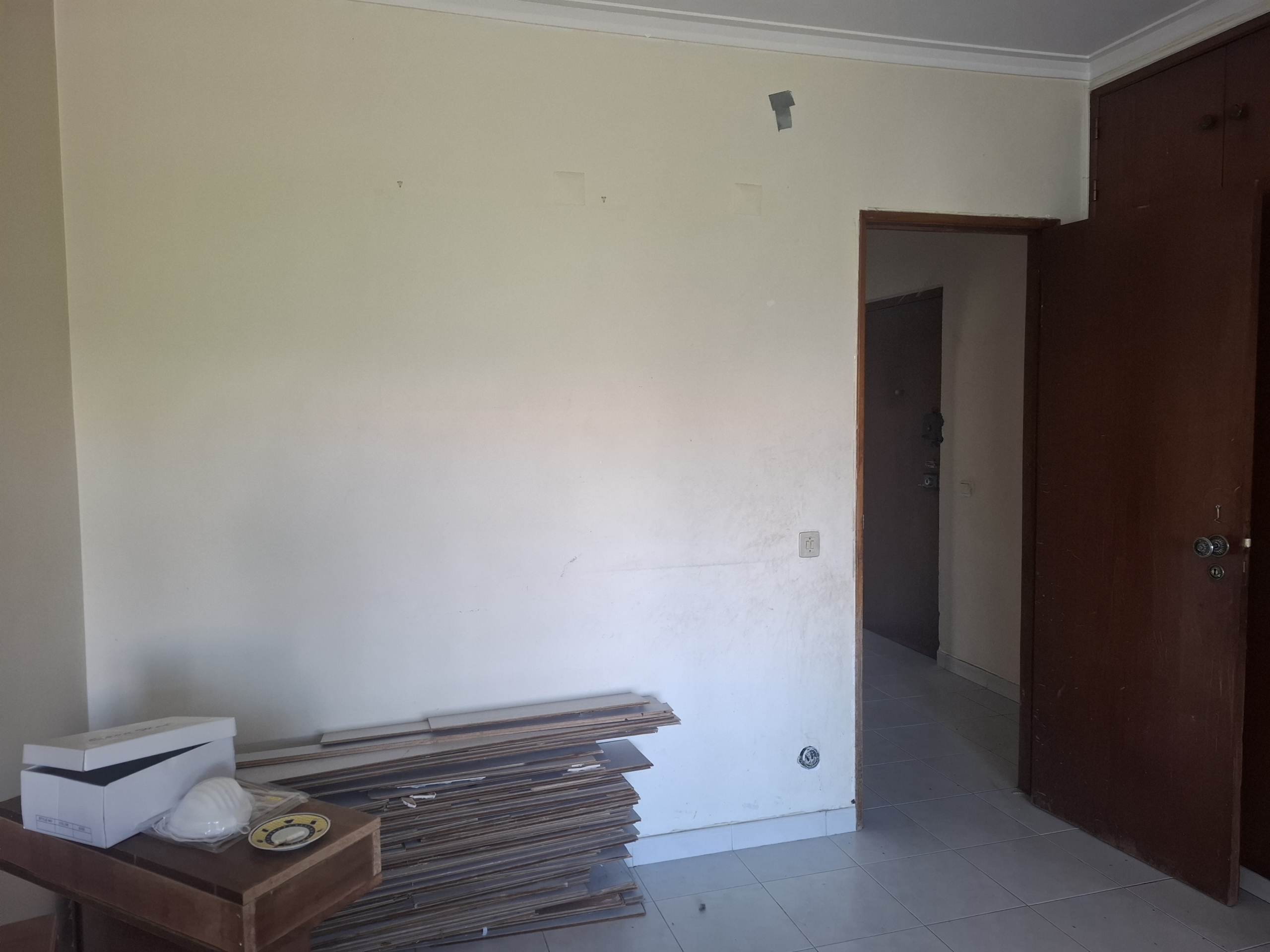 A partially furnished room with a stack of plywood boards along the wall, a brown wooden cabinet door on the right, and a cardboard box in the foreground; walls appear bare with some small patches and a doorway opening visible.