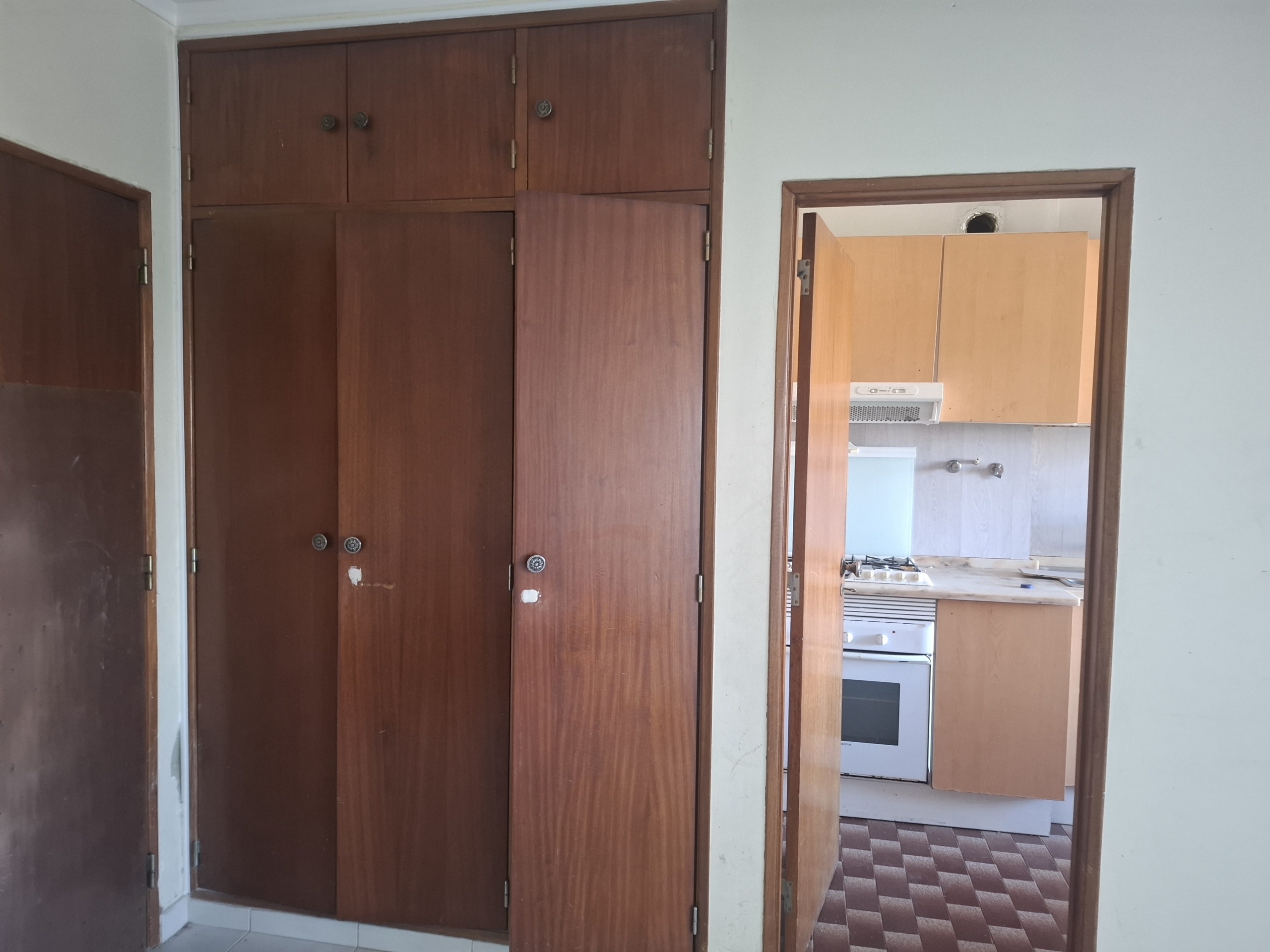 Interior view of a doorway leading to a kitchen; tall wooden wardrobes cover the left wall, with a visible stove oven through an open door on the right.