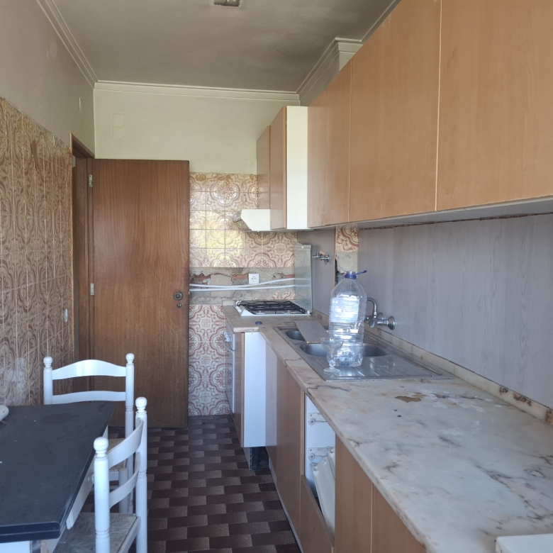 Narrow vintage kitchen with brown cabinets, pink patterned tiles, marble countertops, gas stove, sink, and a small table with white chairs.