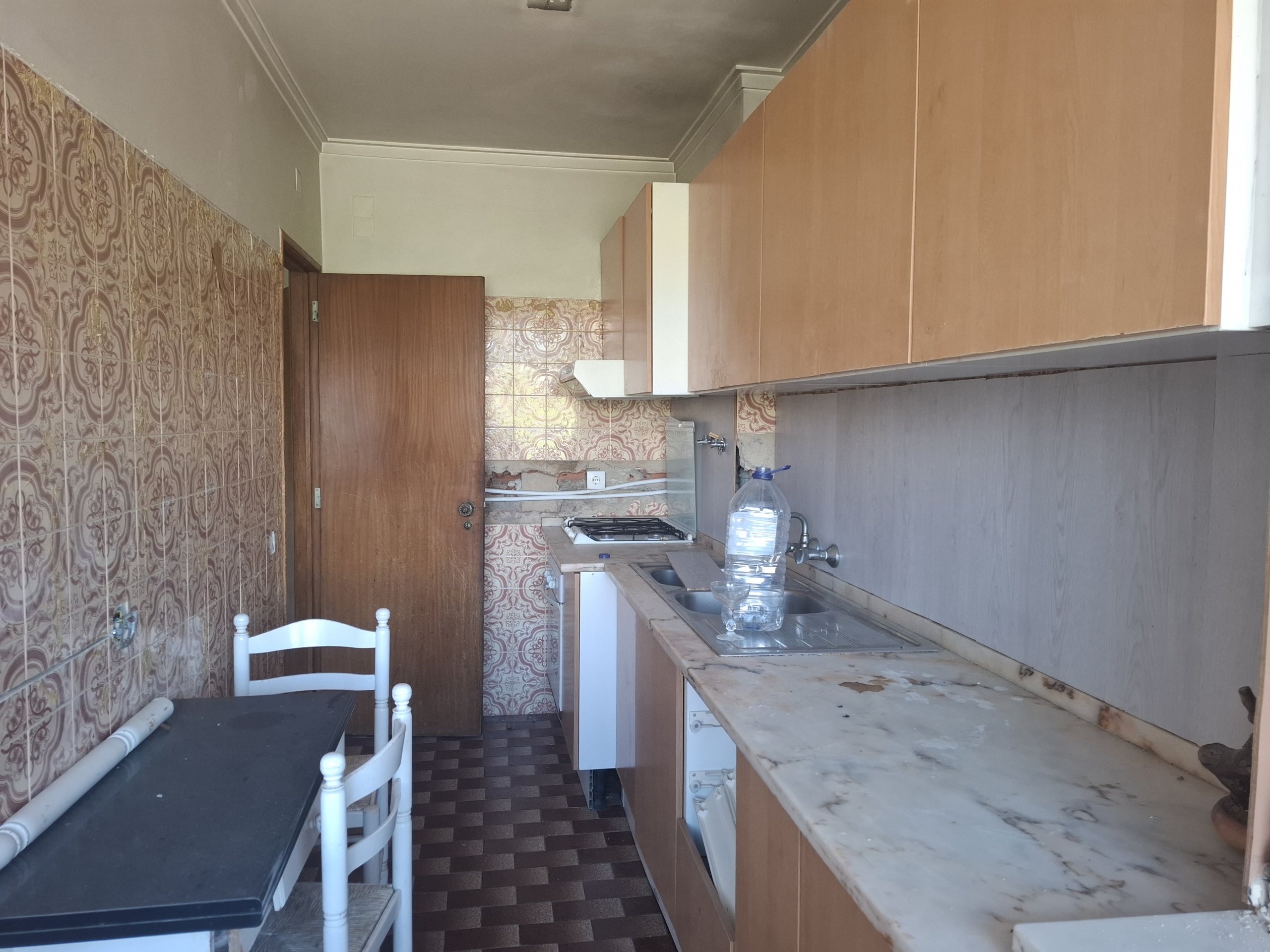 Narrow vintage kitchen with brown cabinets, pink patterned tiles, marble countertops, gas stove, sink, and a small table with white chairs.