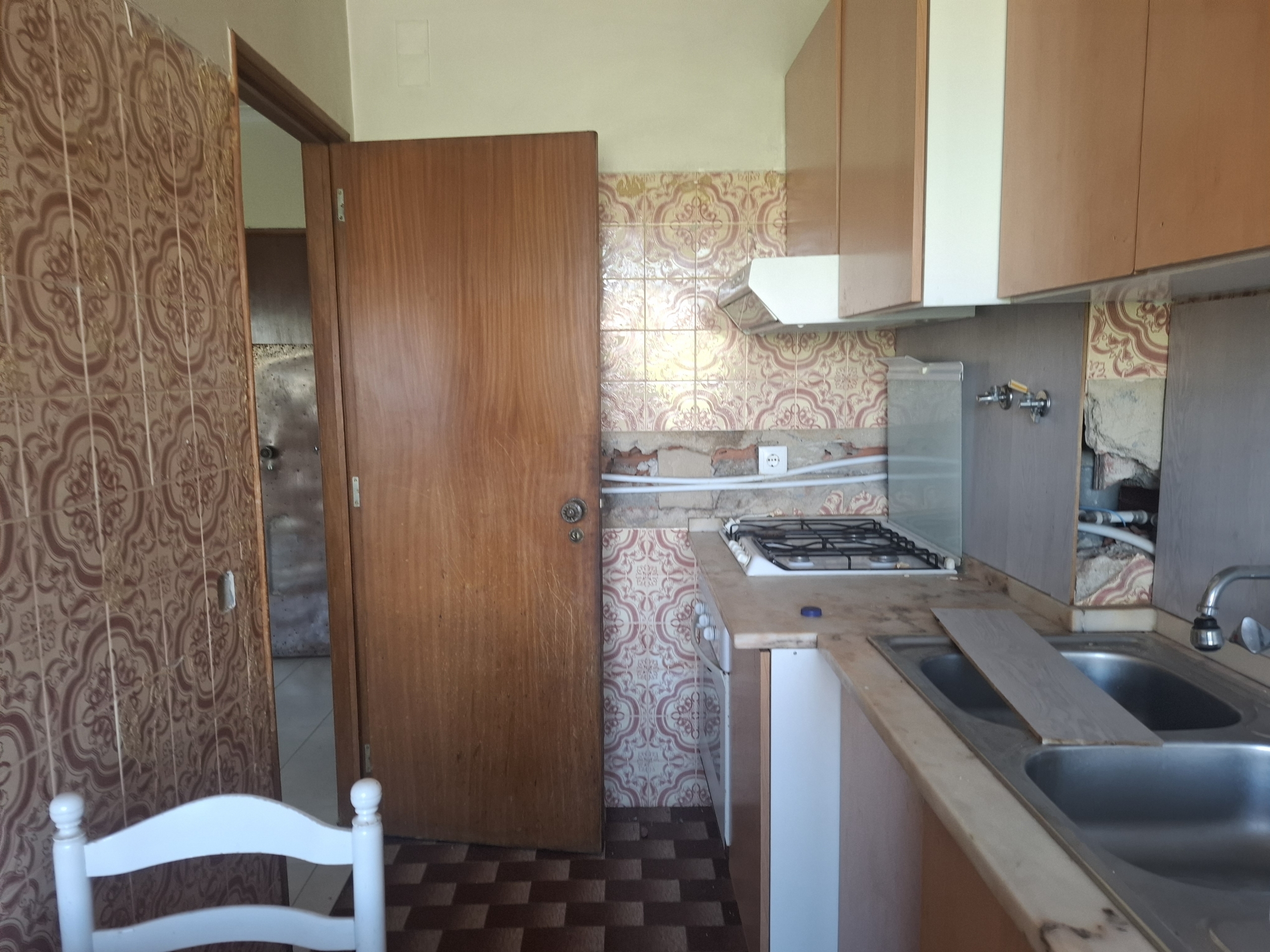 Old kitchen with pink patterned wall tiles, a wooden door ajar, a gas stove, and a double sink visible to the right.