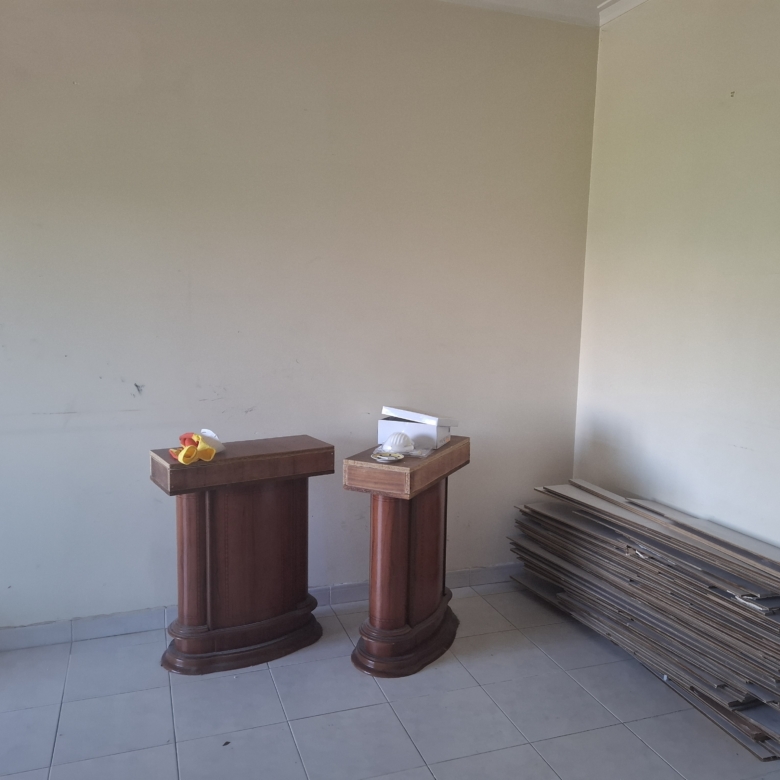 Two dark wooden pedestals in a bare room, with a fabric flower arrangement on the left pedestal and papers on the right pedestal; stacked boards along the wall to the right.