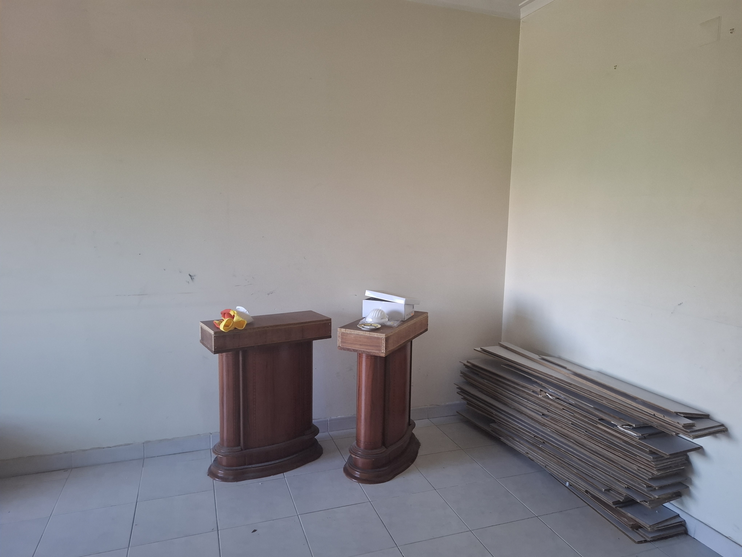Two dark wooden pedestals in a bare room, with a fabric flower arrangement on the left pedestal and papers on the right pedestal; stacked boards along the wall to the right.