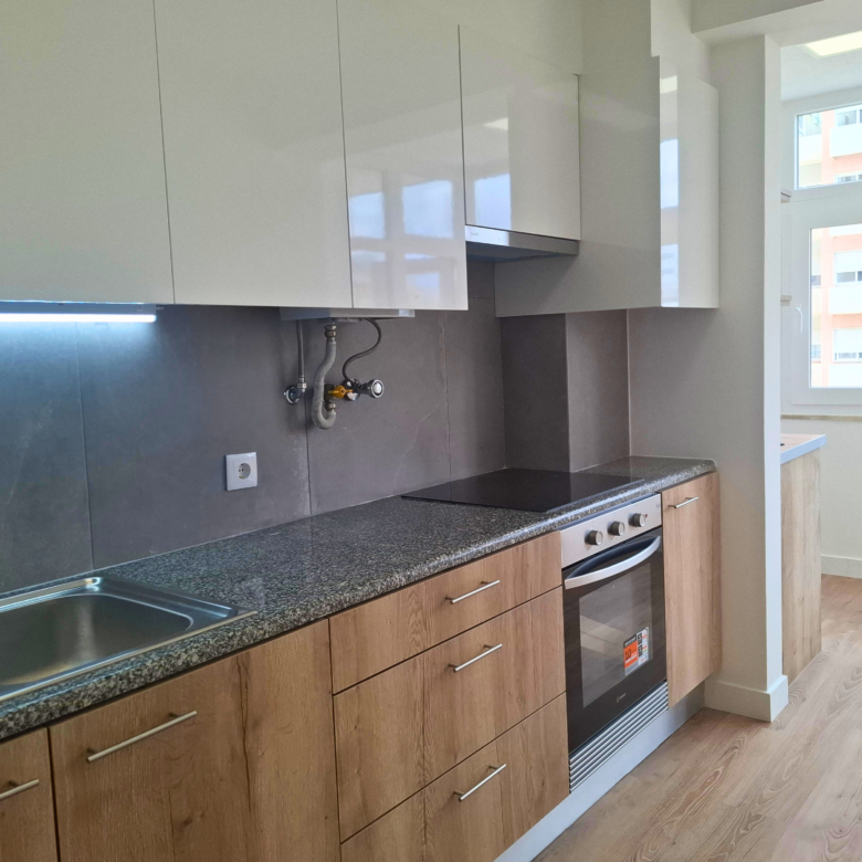 Modern kitchen with white upper cabinets, wood lower cabinets, gray countertop, and stainless steel sink. A windowed wall is visible to the right.