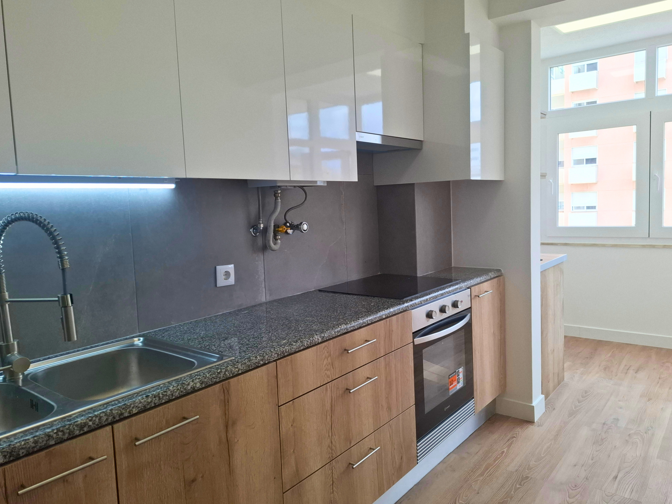 Modern kitchen with white upper cabinets, wood lower cabinets, gray countertop, and stainless steel sink. A windowed wall is visible to the right.