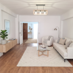 Bright living room with white walls, beige sofa, glass coffee table, and a potted plant; open doorway to corridor.