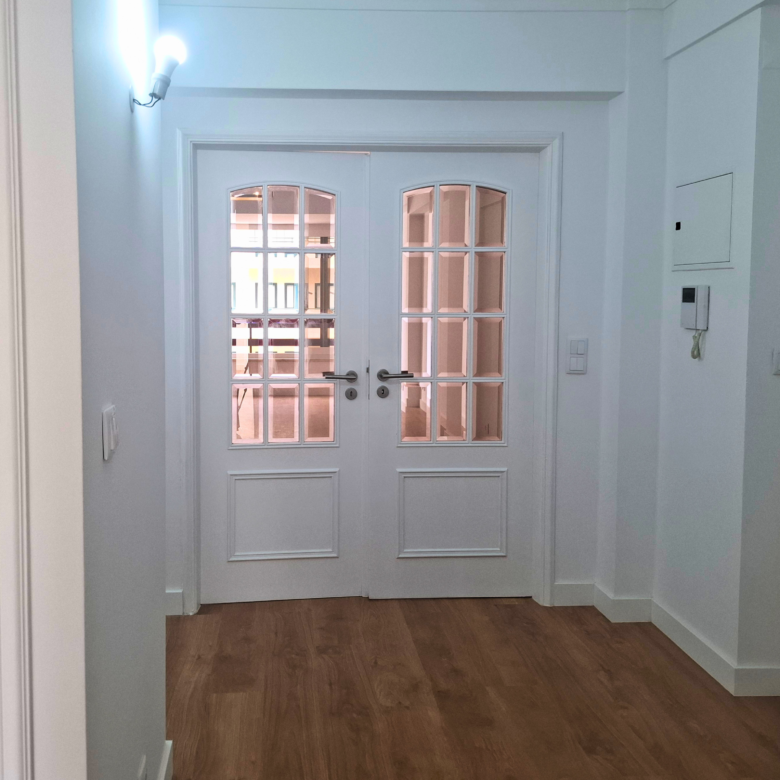 White interior hallway leading to double glass-pane doors, with a hardwood floor and a wall-mounted light fixture on the left.