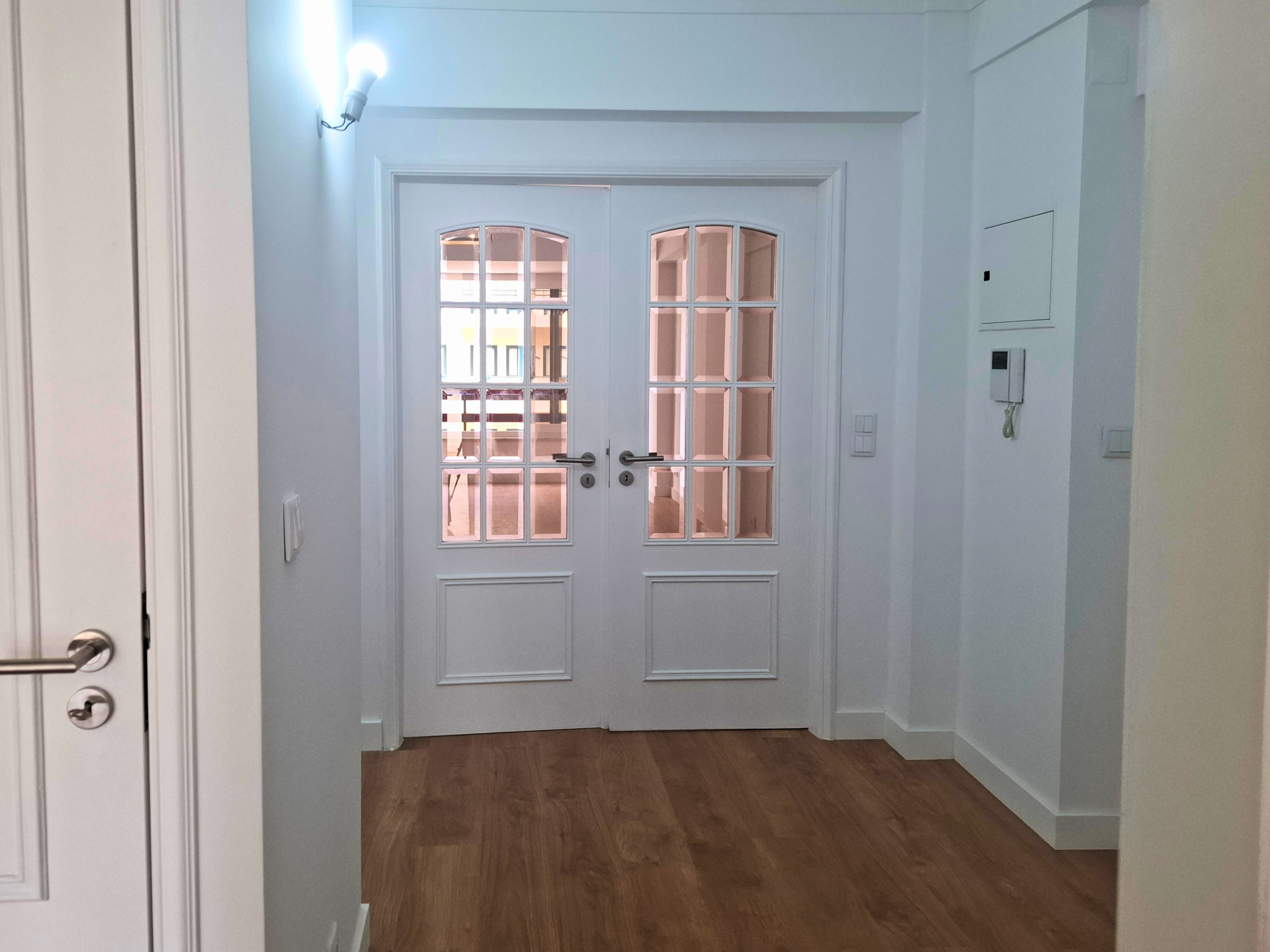 White interior hallway leading to double glass-pane doors, with a hardwood floor and a wall-mounted light fixture on the left.