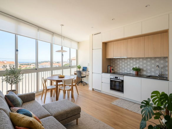 Bright open-plan living area with a white kitchen, wooden accents, and a round dining table by wide windows showing a city view.