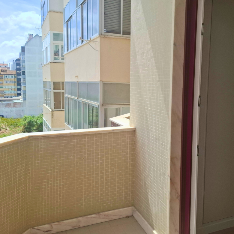 Balcony corner with beige tiled walls and a glass door with a purple frame; city buildings and a blue sky in the distance.