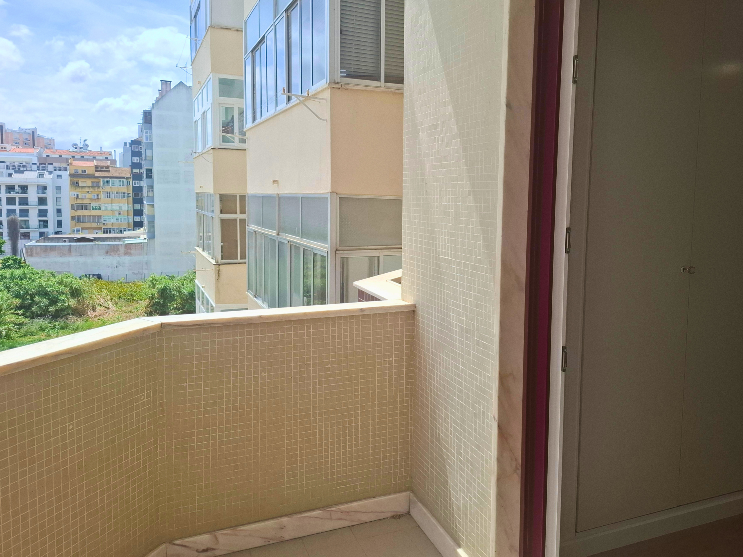 Balcony corner with beige tiled walls and a glass door with a purple frame; city buildings and a blue sky in the distance.