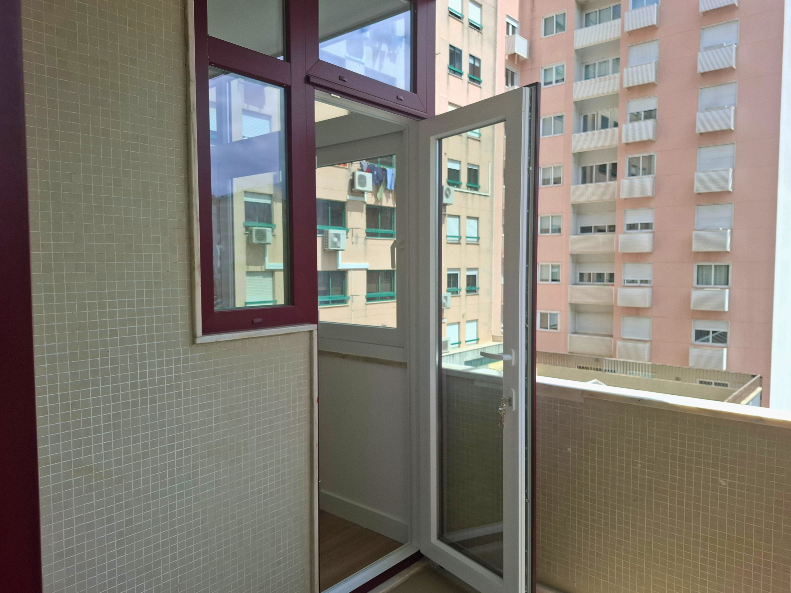 Open glass door to a tiled balcony, showing an urban building with windows and visible air conditioning units outside.