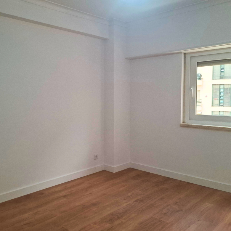 Empty white room corner with a large window on the right and wood flooring beneath.