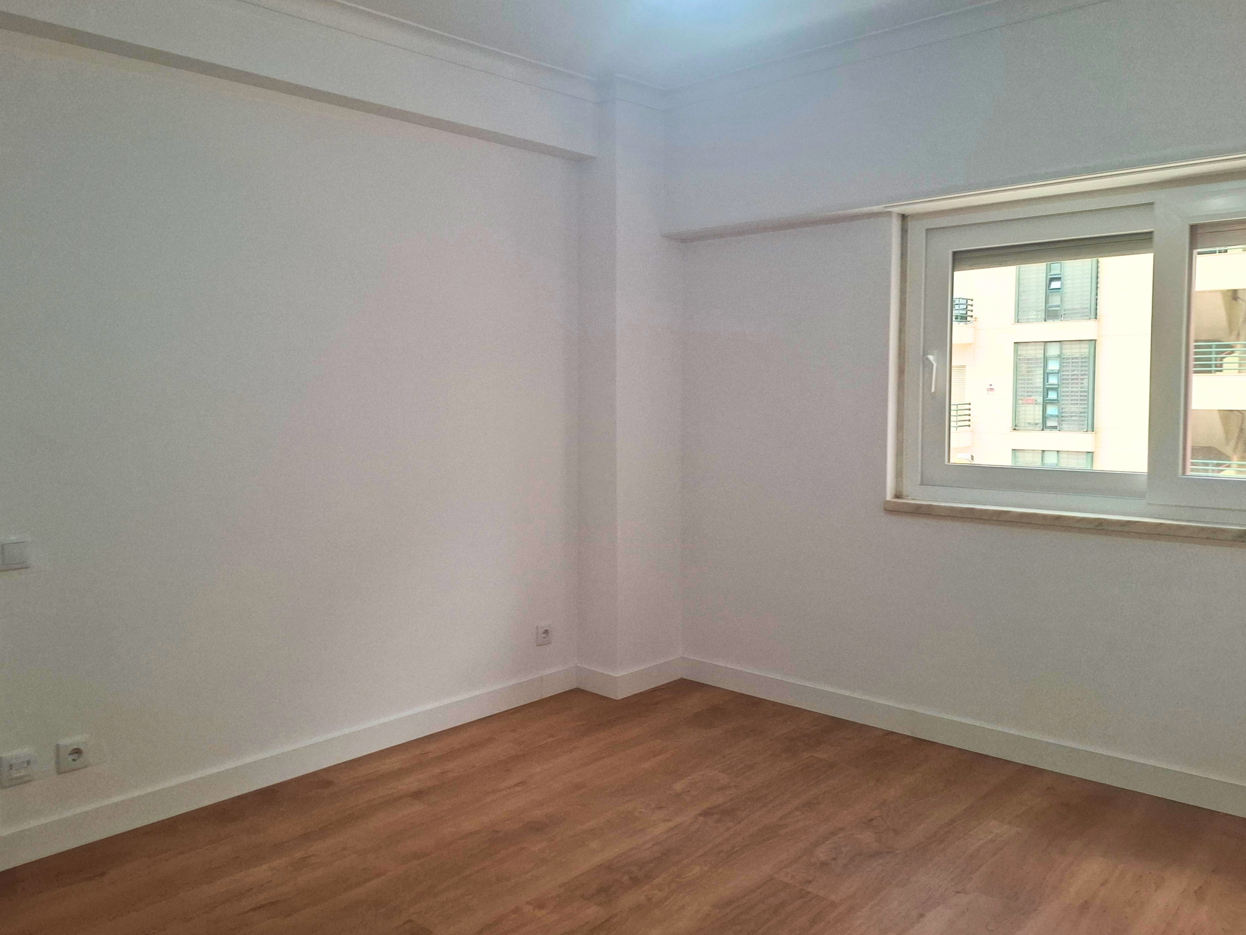 Empty white room corner with a large window on the right and wood flooring beneath.