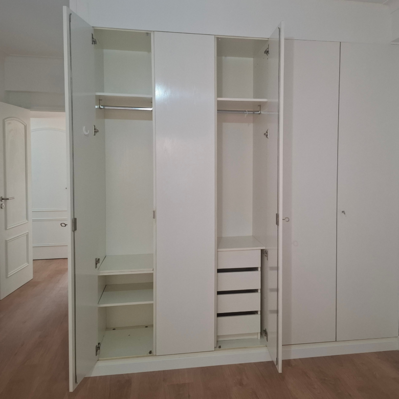 Open white wardrobe with multiple shelves and hanging rail, showing drawers at the bottom in a bright room.