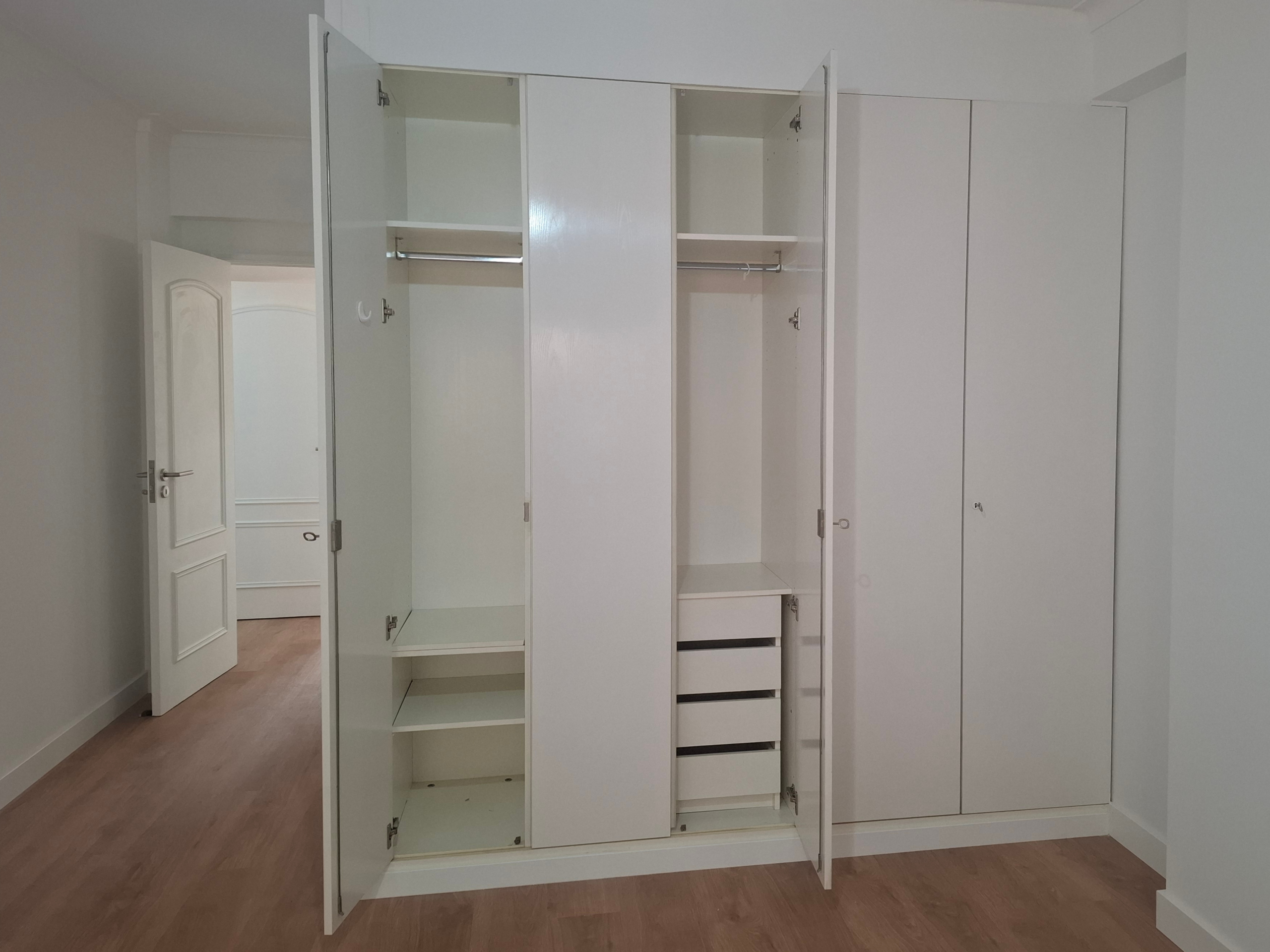 Open white wardrobe with multiple shelves and hanging rail, showing drawers at the bottom in a bright room.