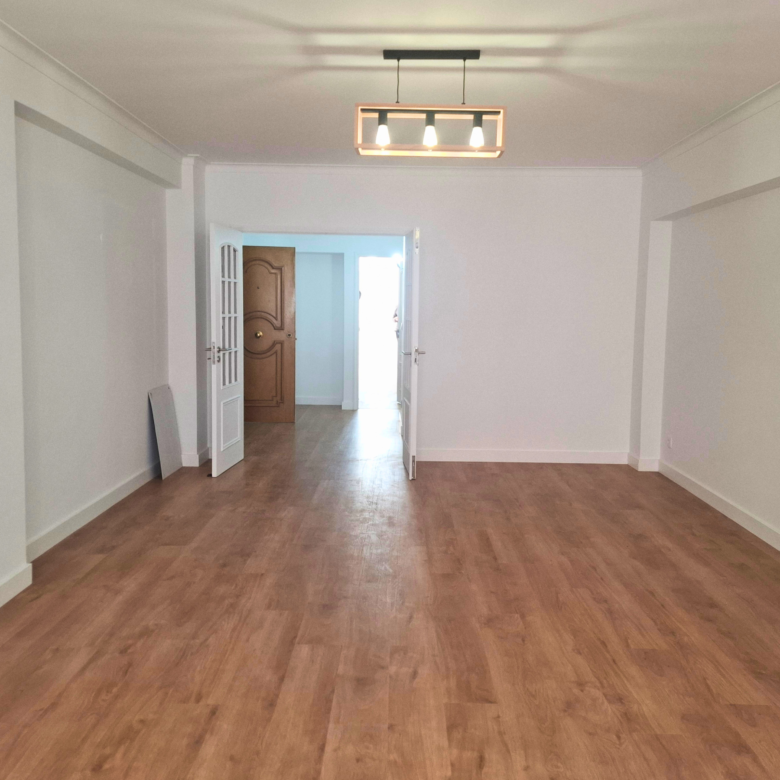 Empty, spacious room with light wood floors and white walls; double doors open toward a sunlit area, and a modern rectangular ceiling light hangs overhead.