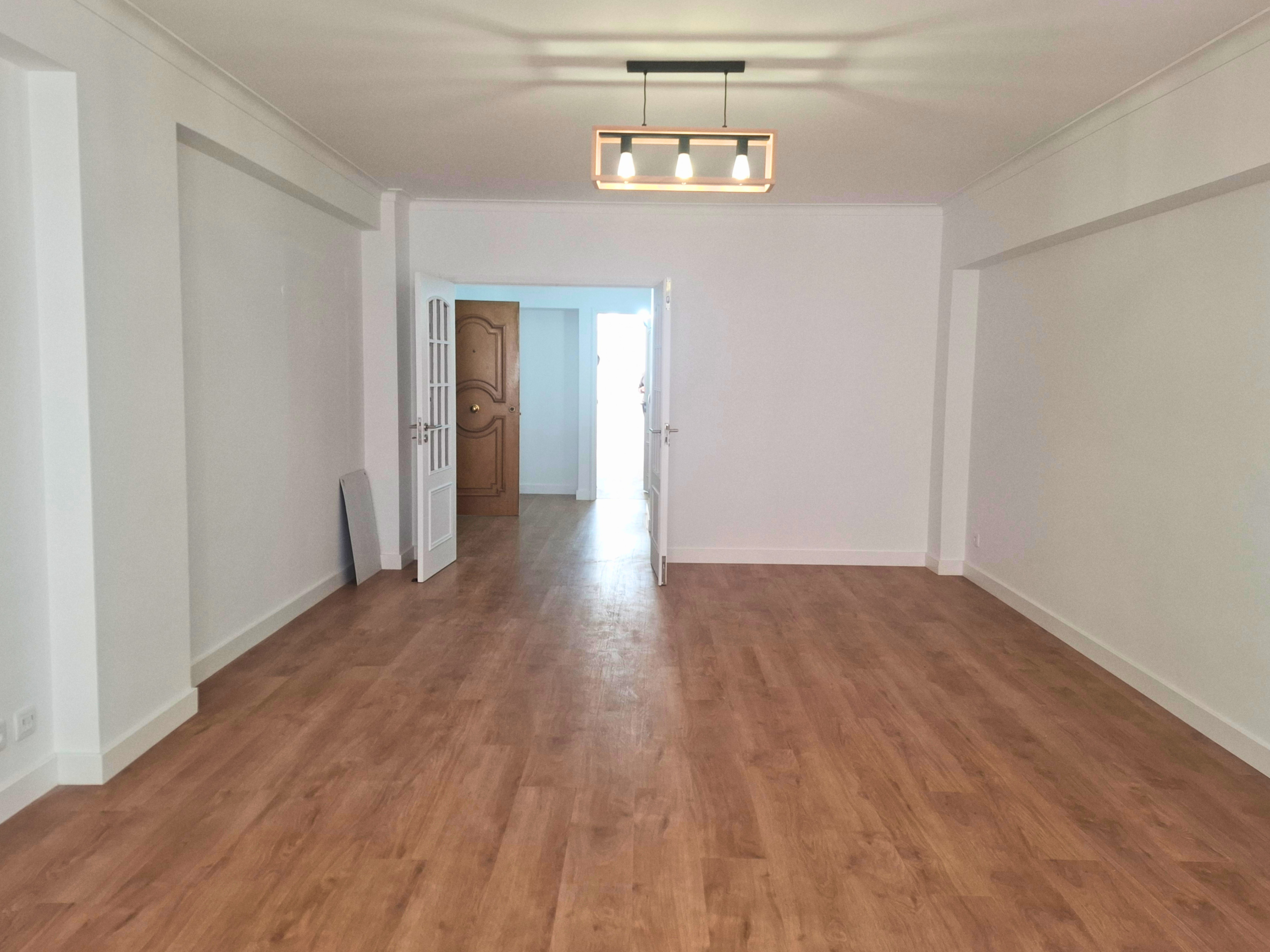 Empty, spacious room with light wood floors and white walls; double doors open toward a sunlit area, and a modern rectangular ceiling light hangs overhead.